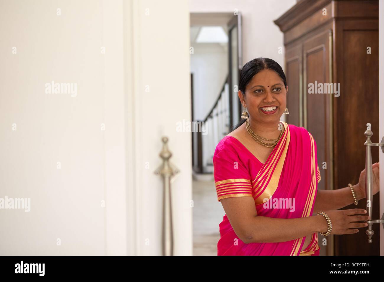 Lächelnde indianerin in rosa goldenem Saree und öffnet hölzerne Schranktür im Flur Stockfoto