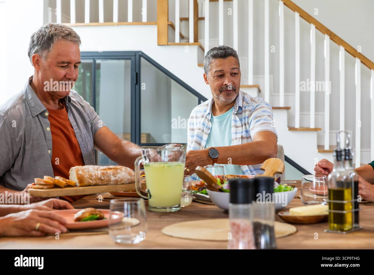 Ältere Freunde teilen ihre Mahlzeit im Speisesaal zu Hause mit Schneidebrett, Salatschüssel, Limonadenkrug Stockfoto