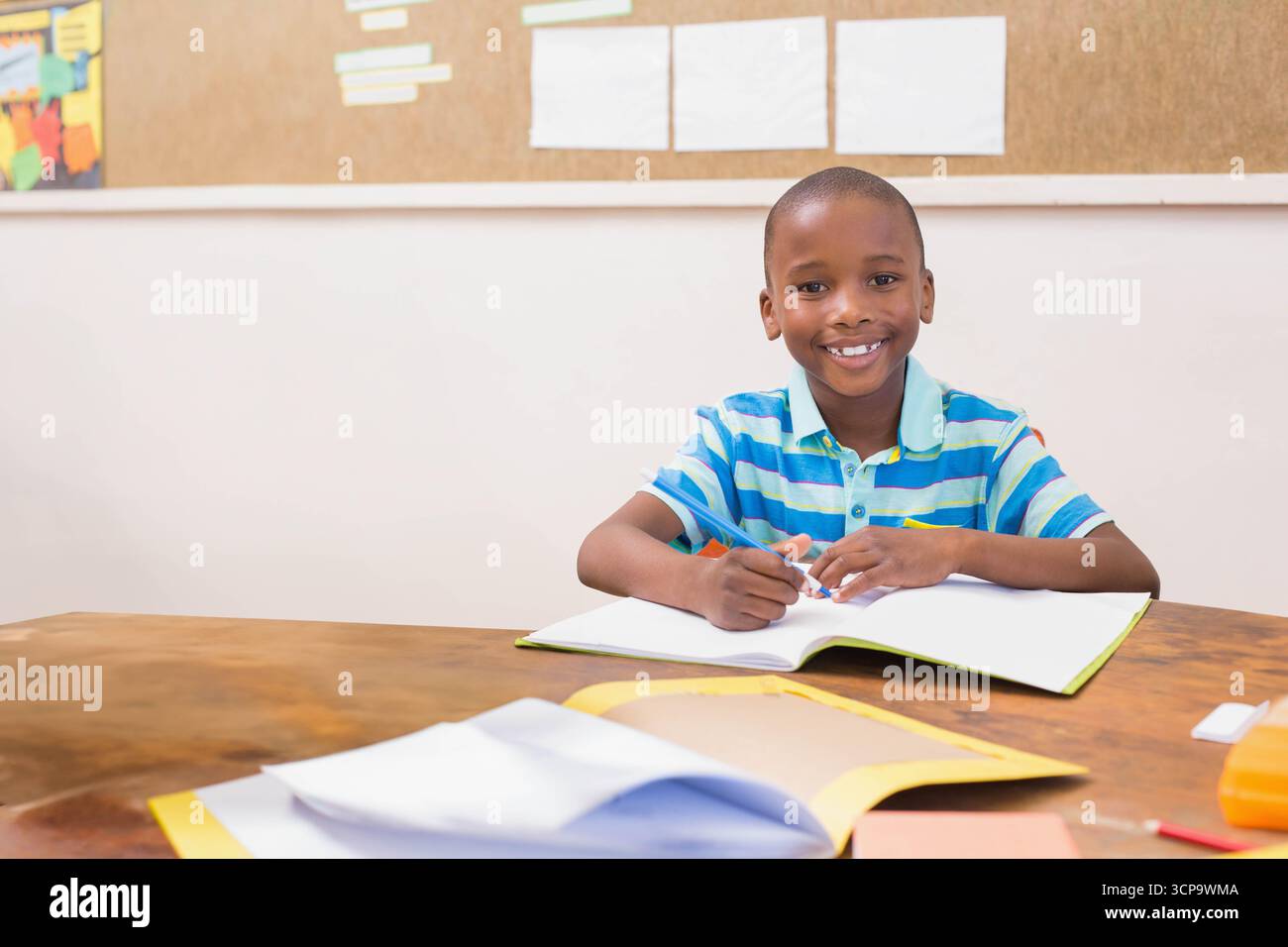 Ein afroamerikanischer Junge lächelt in die Kamera, während er den Bleistift über ein offenes Notizbuch hält Stockfoto