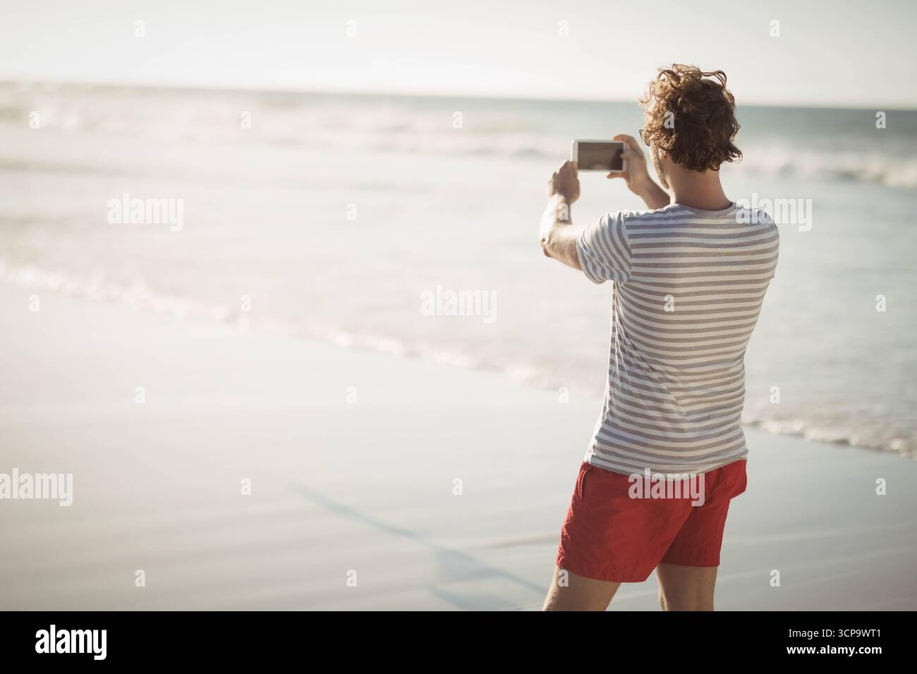 Mann in gestreiftem Hemd, roten Badeshorts, die den Horizont des Meeres auf dem Smartphone am Strand einfangen, Kopierraum Stockfoto