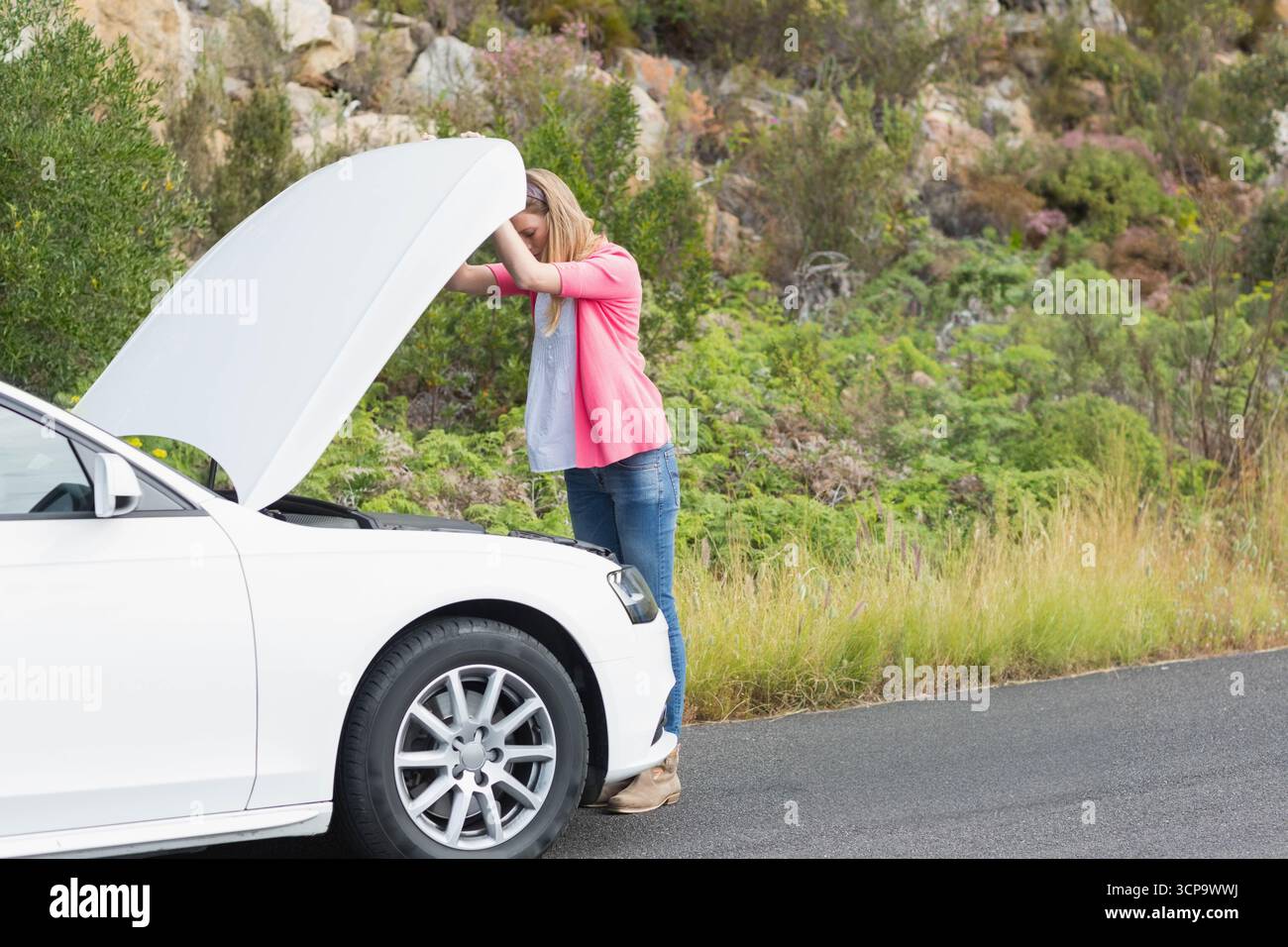 Frau, die den Motorraum unter der erhöhten Haube einer weißen Limousine neben einer asphaltierten Landstraße inspiziert Stockfoto