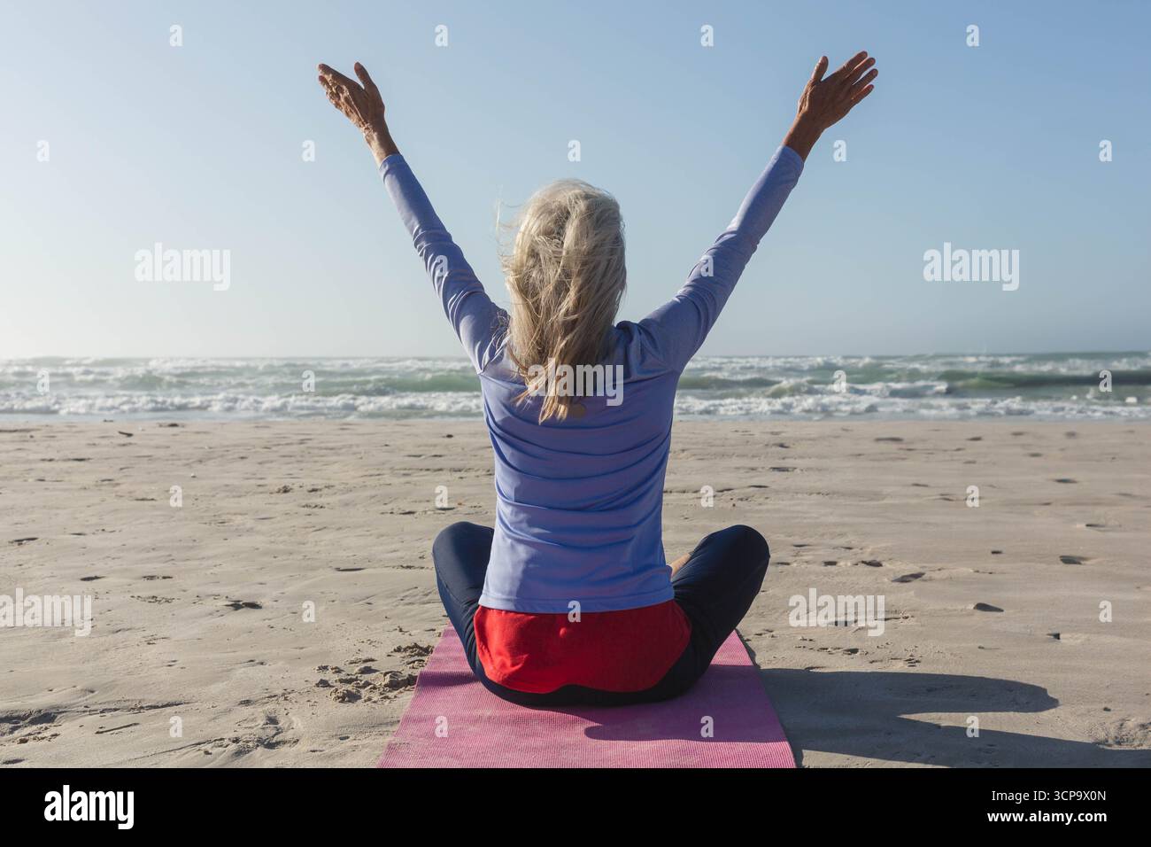 Frau, die Yoga übt, sitzend im Kreuz auf rosa Matte am Meer in sportlicher Kleidung Stockfoto