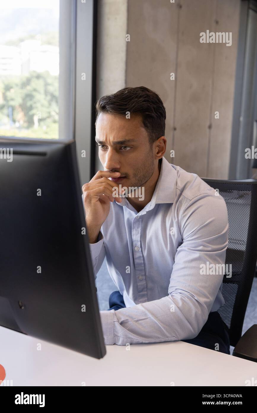 Mann in blauem Hemd, der sich vor den Schreibtisch lehnt und auf den Computermonitor im Büro starrt Stockfoto