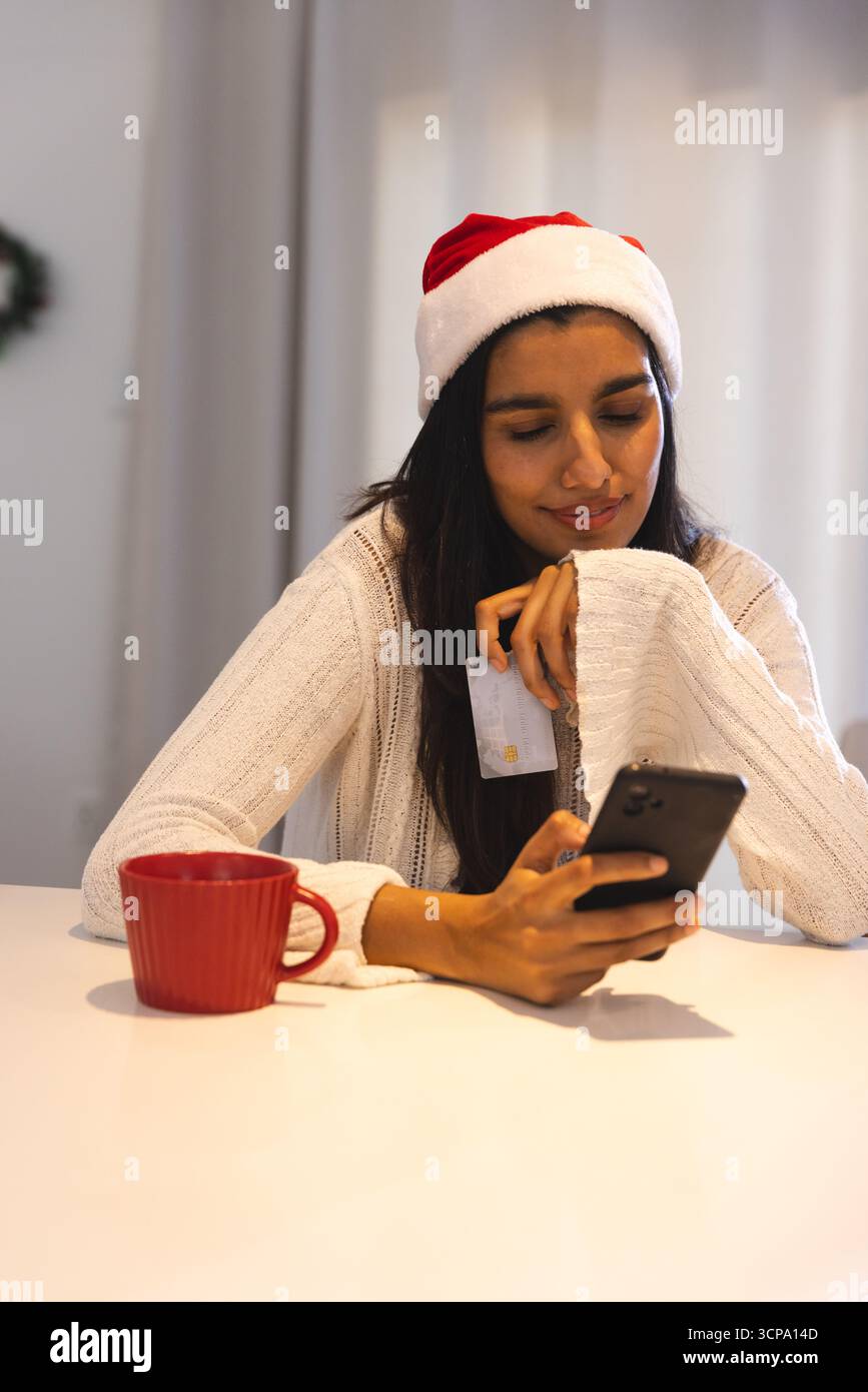 Frau mit Weihnachtsmann-Pullover und -Hut, mit Kreditkarte, mit Smartphone am Tisch mit Becher Stockfoto