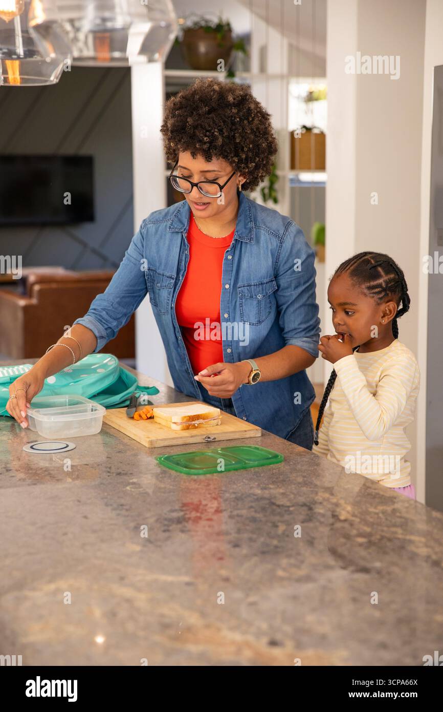 Vielfältige Mutter mit Tochter, die Sandwich mit Karotten zubereitete, Behälterdeckel auf Kücheninsel Stockfoto
