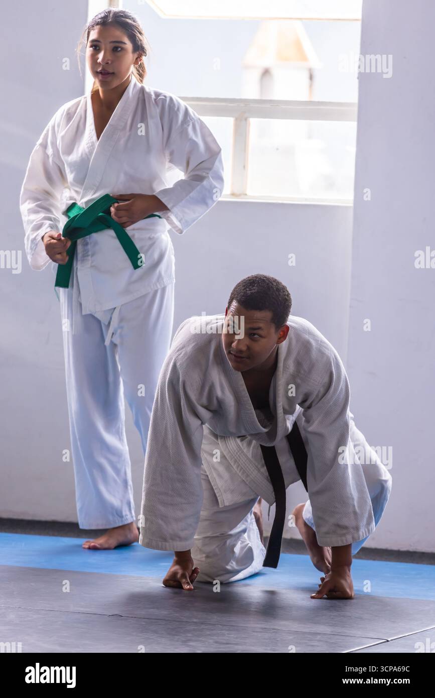 Asiatischer Teenager, der den grünen Gürtel auf Gi anpasst, als afroamerikanischer Ausbilder, der auf der Matte kniet Stockfoto
