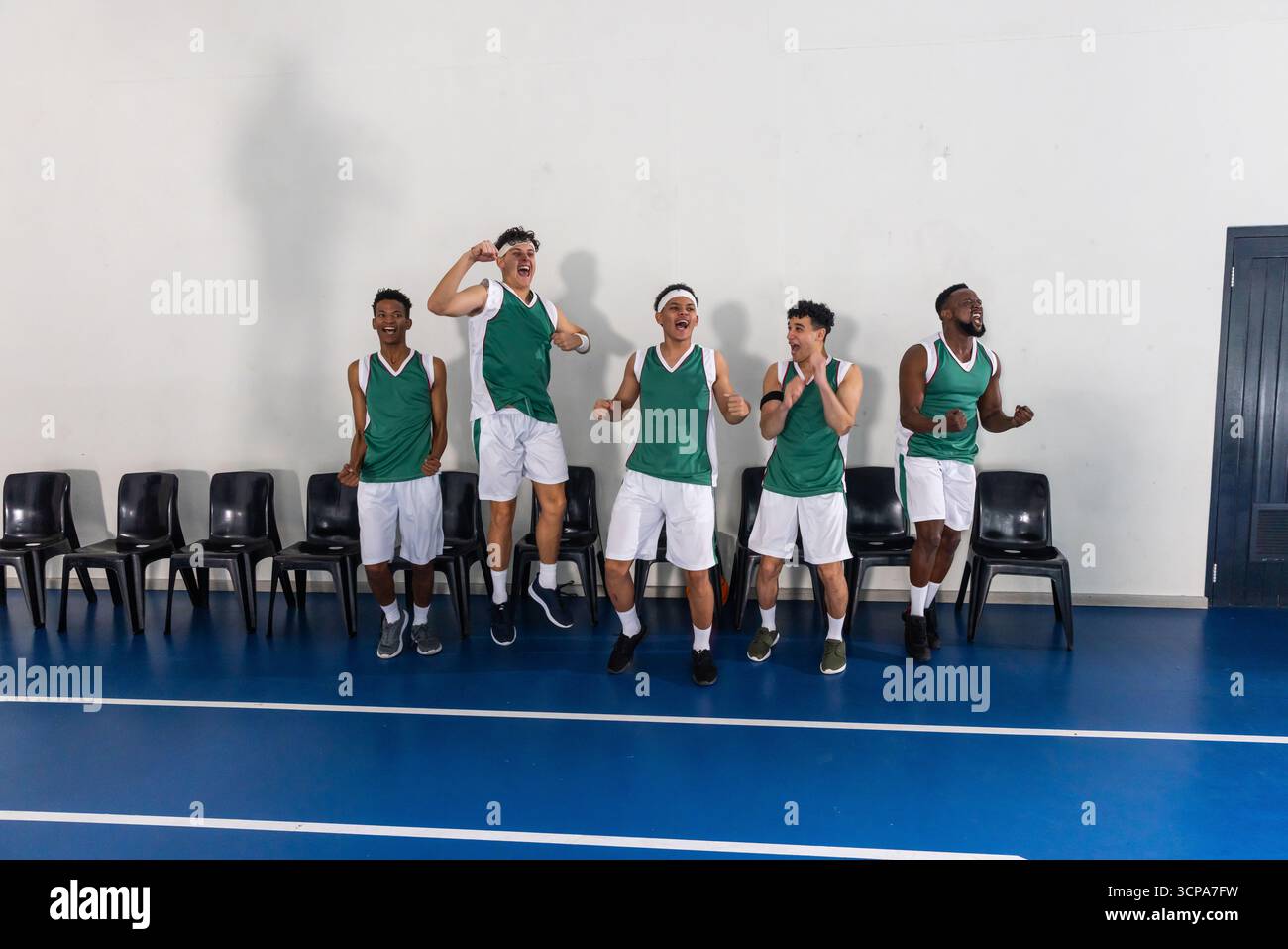 Verschiedene männliche Basketballkollegen in grünen Trikots feiern auf dem blauen Platz in der Nähe von Stühlen Stockfoto