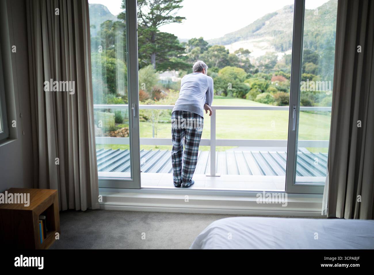 Ein älterer Mann im Pyjama, der sich auf einem Balkongeländer stützt und durch Glastüren auf den gepflegten Rasen blickt Stockfoto