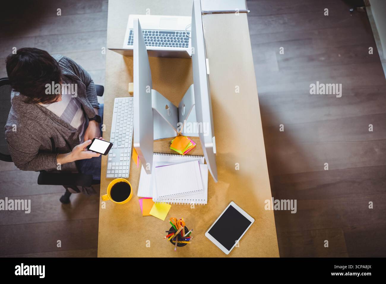 Männlicher Büroangestellter, der das Smartphone überprüft, während er am Bürotisch mit Monitoren sitzt, Kopierraum Stockfoto