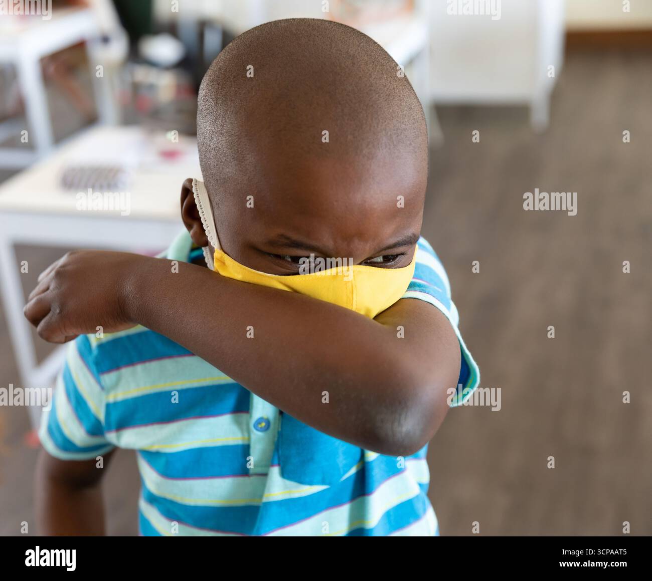 Der afroamerikanische Junge hustet in den Ellenbogen, trägt gelbe Gesichtsmaske im Klassenzimmer mit weißen Tischen Stockfoto