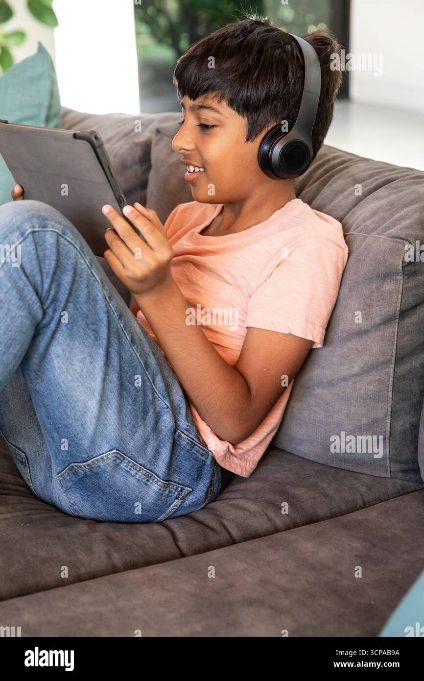 südasiatischer Schuljunge, der im Wohnzimmer auf einem grauen Sofa sitzt und ein Tablet mit Kopfhörern trägt Stockfoto