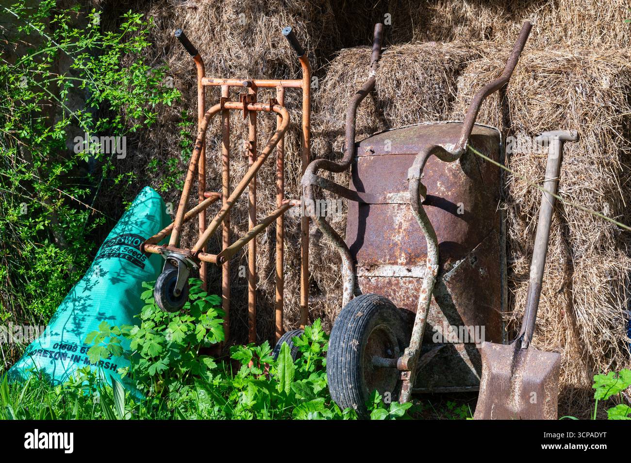 Rostige Schubkarre, Sackkarre und Schaufel auf Heuballen in einem traditionellen Bauernhof mit Frühlingsgrün im Vordergrund Stockfoto