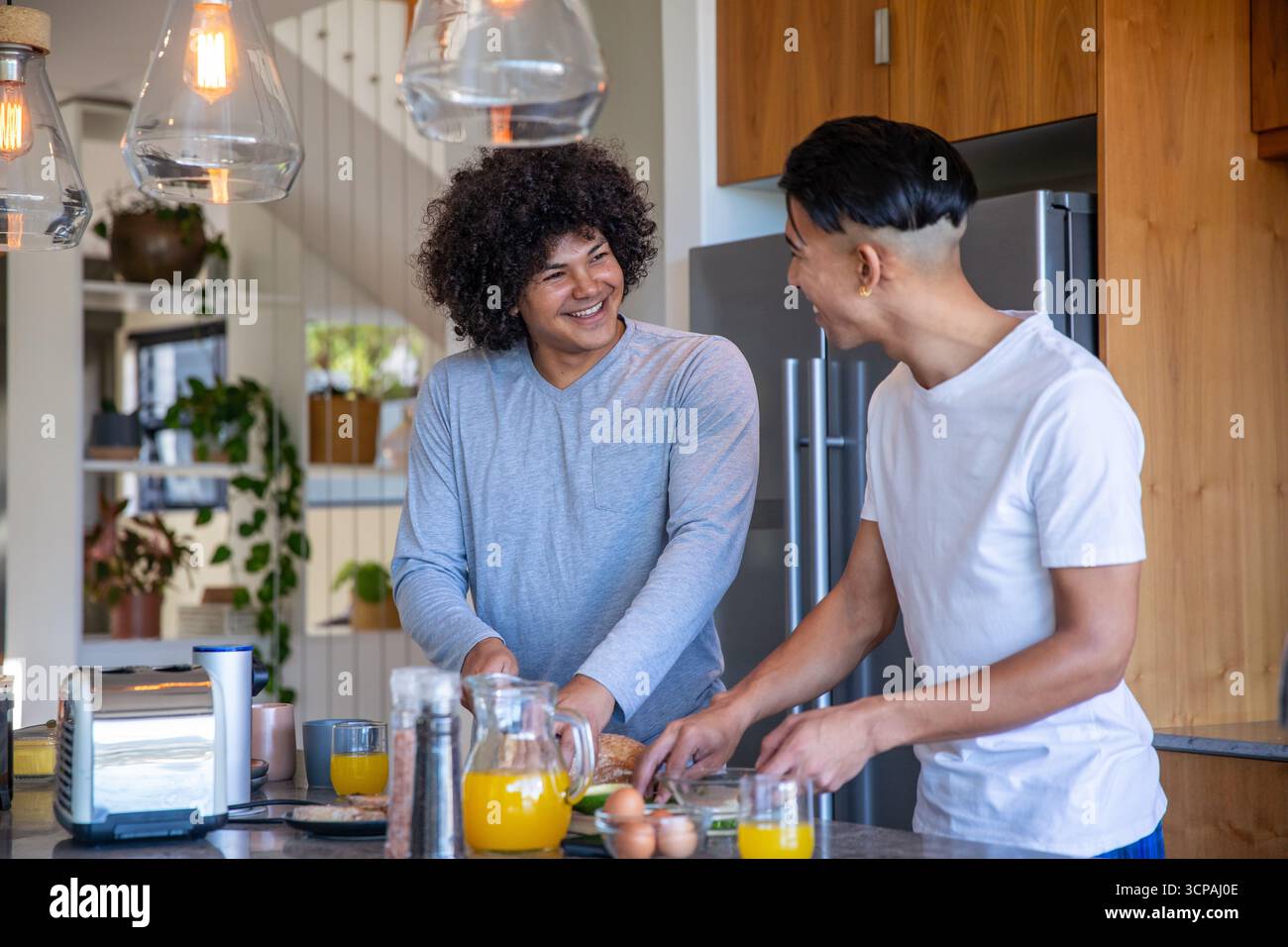 Männliches Paar, das Frühstück auf der Kücheninsel zubereitete und Brot an Bord mit Toaster und Saft schneidet Stockfoto