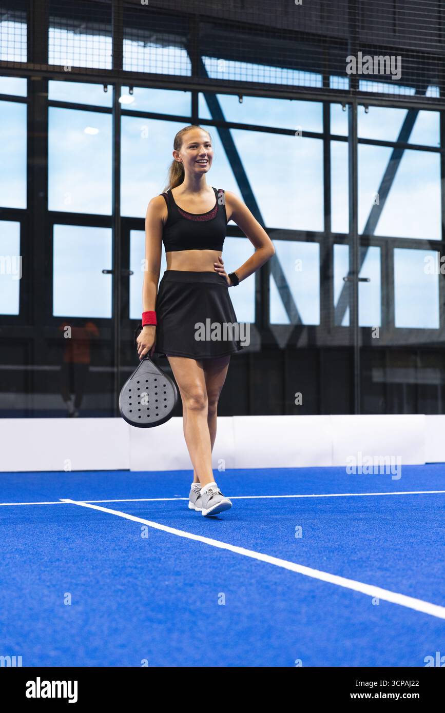 Weibliche Athletin, die auf dem blauen Padel-Tennisplatz steht und einen Padel-Schläger hält, lächelt mit Glaswänden Stockfoto