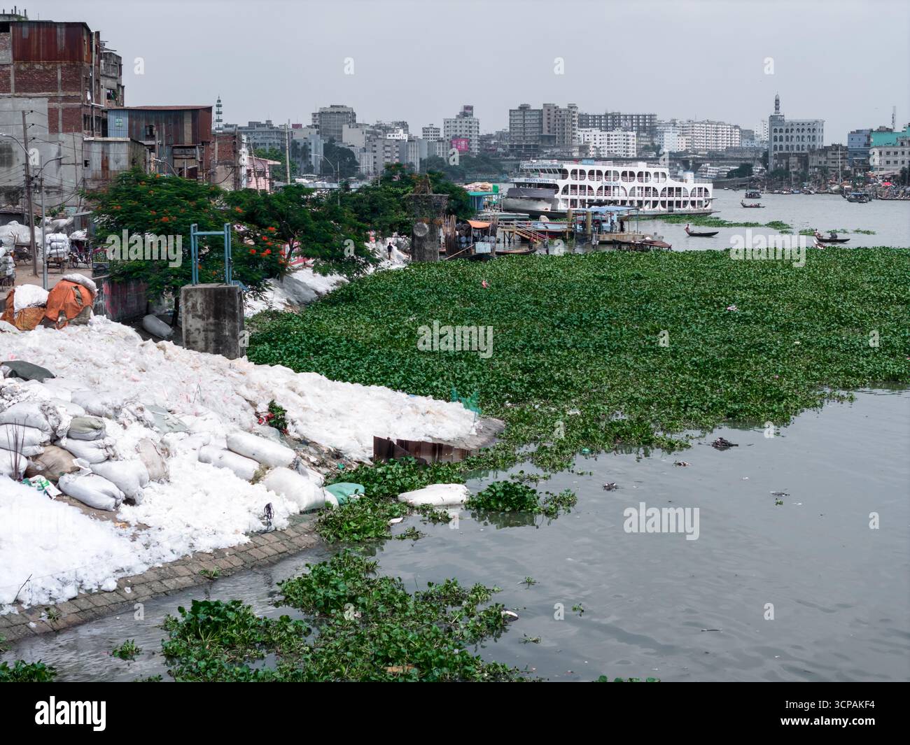 Dhaka, Bangladesch - 10. September 2025: Aus der Vogelperspektive auf den Buriganga River zeigt sich ein starker Kontrast zwischen der Skyline der Stadt und den mit Wasserhyazinthen bedeckten Ufern. Stockfoto