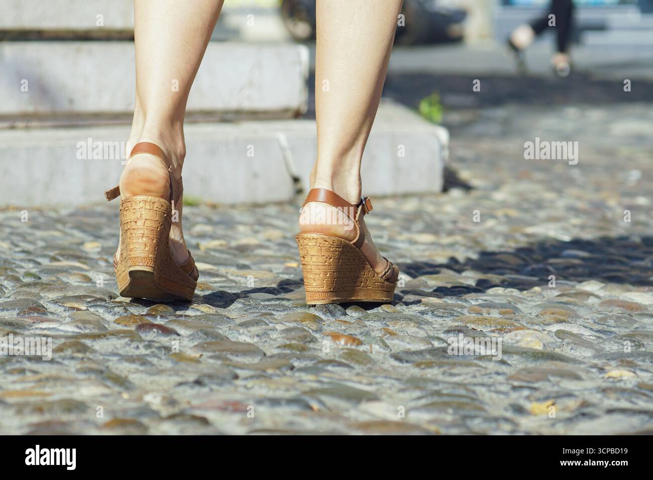 Weibliche Füße in Keilschuhen, die auf Steinpflaster treten. Symbolisiert Sommermode, langsamen Tourismus, achtsames Reisen, urbanen Lebensstil, historische Straßen Stockfoto