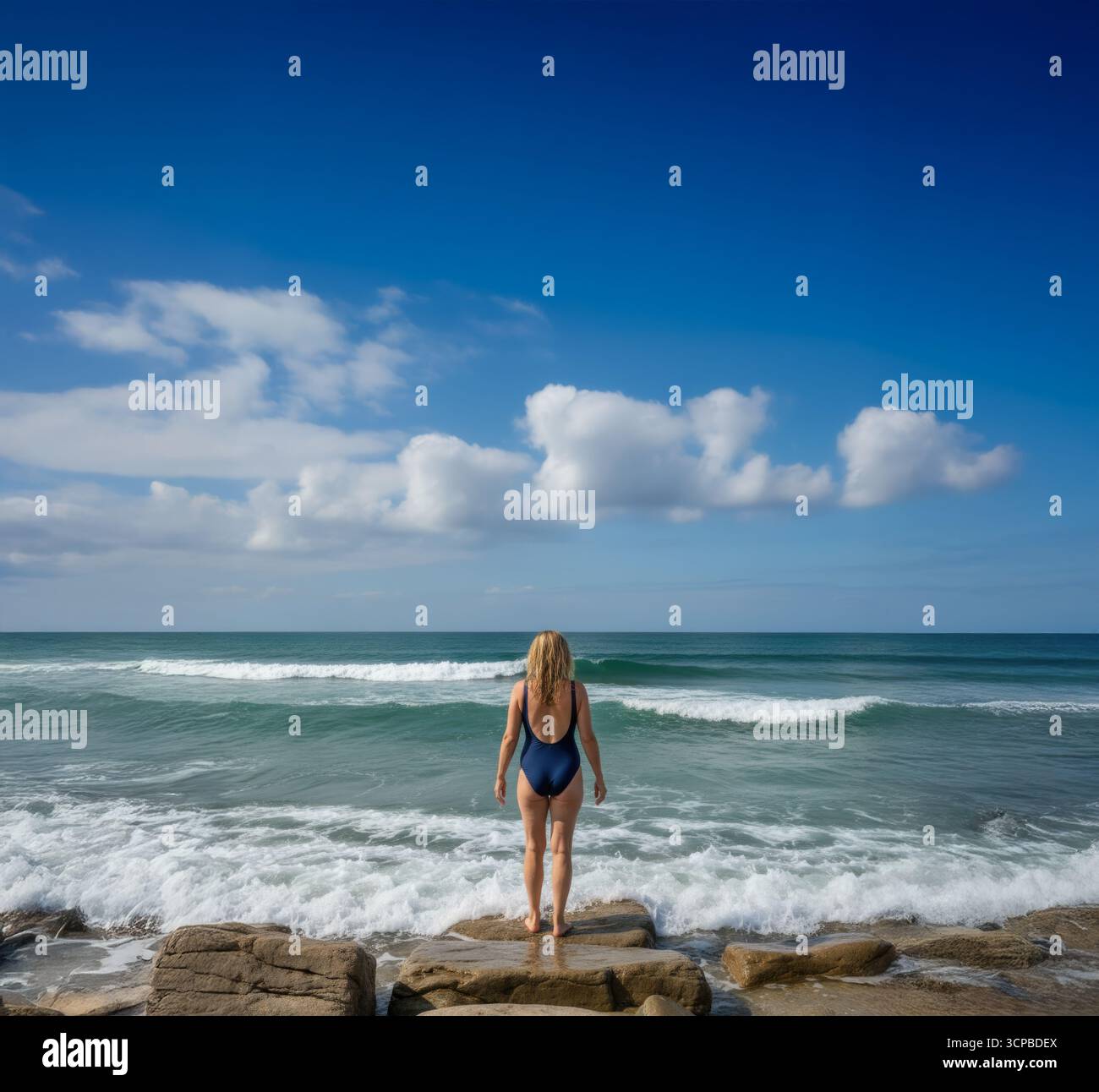 Eine Frau im Badeanzug steht auf Felsen und betrachtet die Wellen des Ozeans, bevor sie an einem schönen sonnigen Tag ein erfrischendes Bad in der Wildnis nimmt. Stockfoto