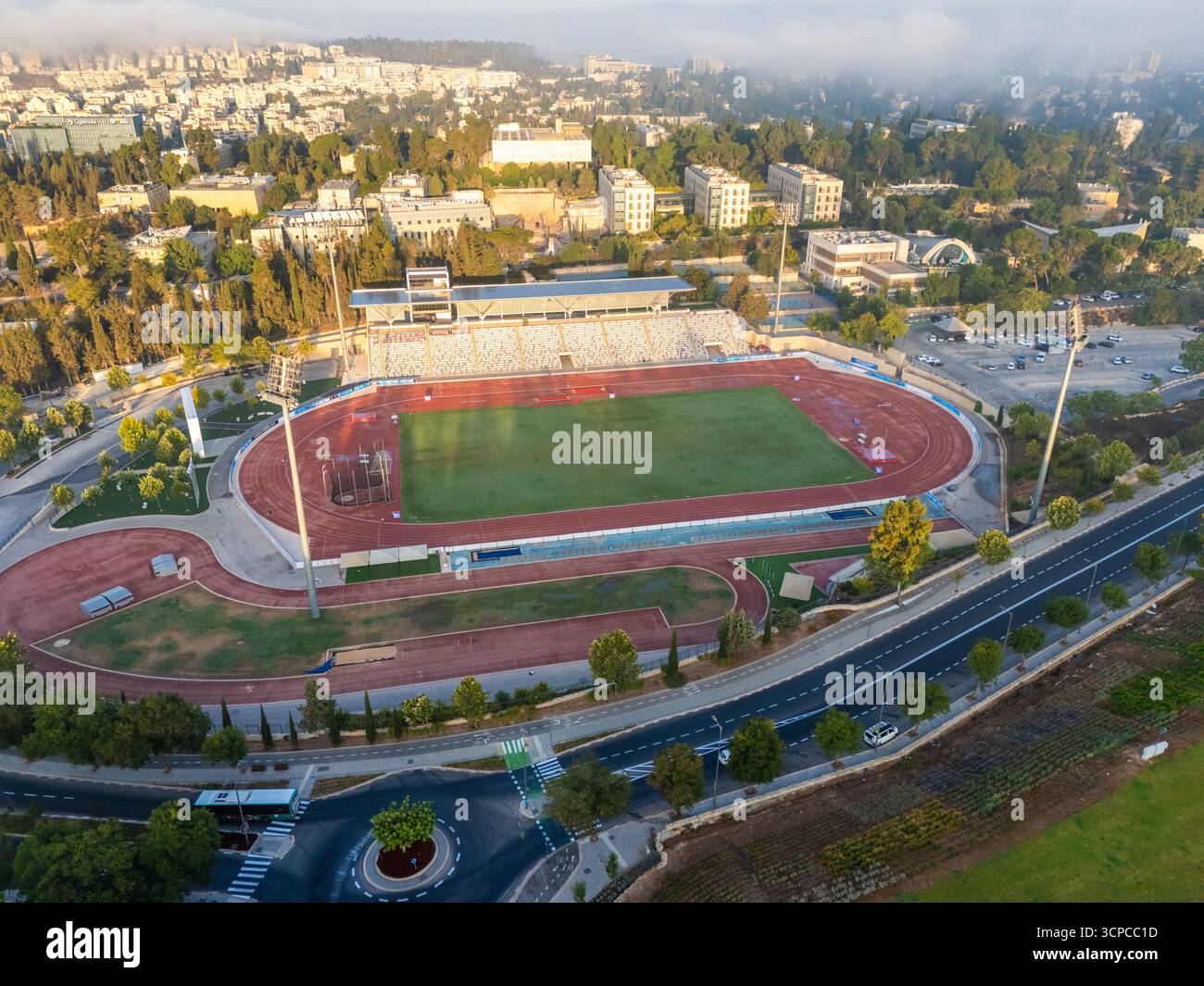 Aus der Vogelperspektive auf ein Givat RAM Stadion mit roten Laufbahnen rund um ein pulsierendes grünes Feld, eingebettet in eine weitläufige Stadtlandschaft und üppige Bäume, Jerusalem, Israel. Stockfoto