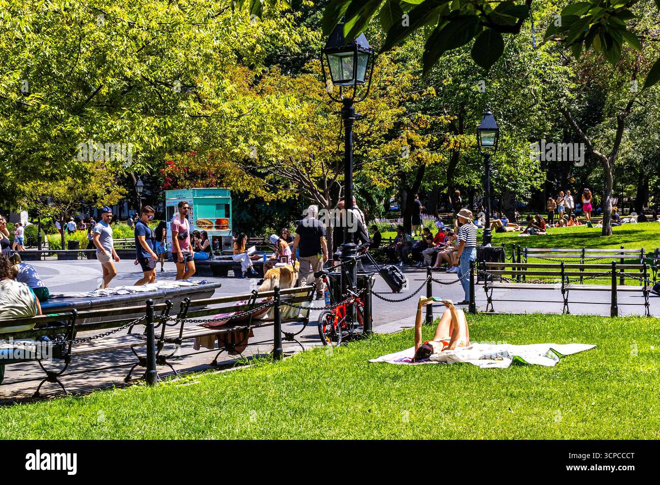 Frau in einem Bikini, die in der Sommersonne bräunt, während sie auf Gras im Washington Square Park, New York City, liest. Menschenmassen genießen warmes Wetter. Stockfoto