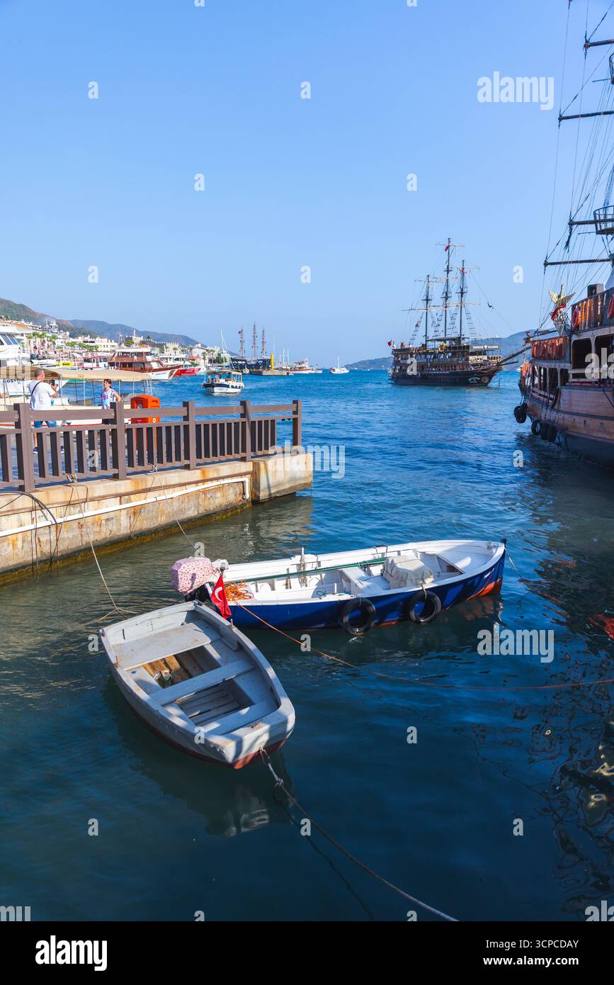 Marmaris, Türkei - 21. August 2024: Ein lebhafter Küstenhafen mit kleinen Booten, die in der Nähe eines Piers angedockt werden, und größeren Segelschiffen, die in der Ferne vor Anker liegen Stockfoto