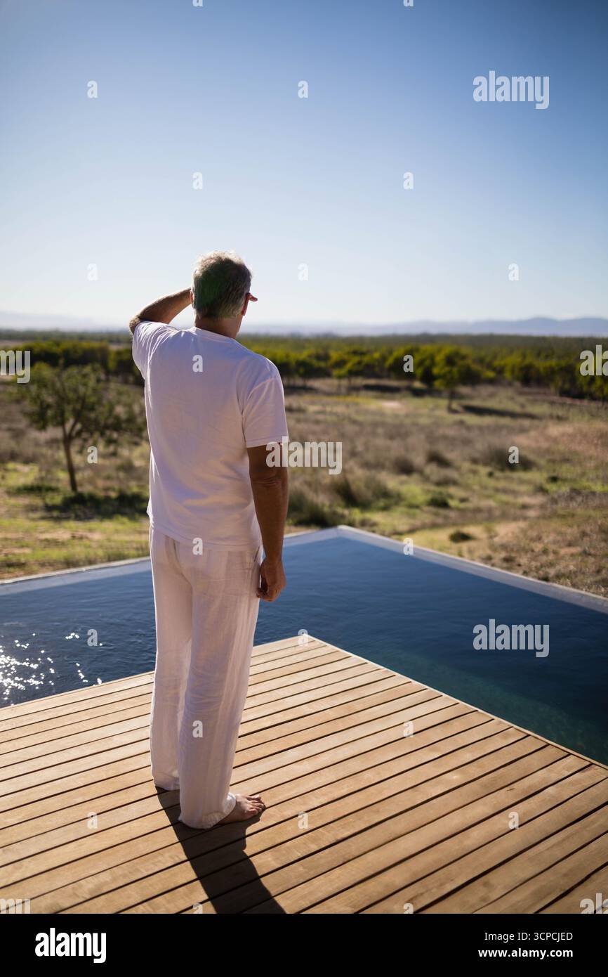 Reifer Mann, der barfuß auf der Holzterrasse am Infinity-Pool steht und auf trockenes Gestrüpp und Berge blickt Stockfoto