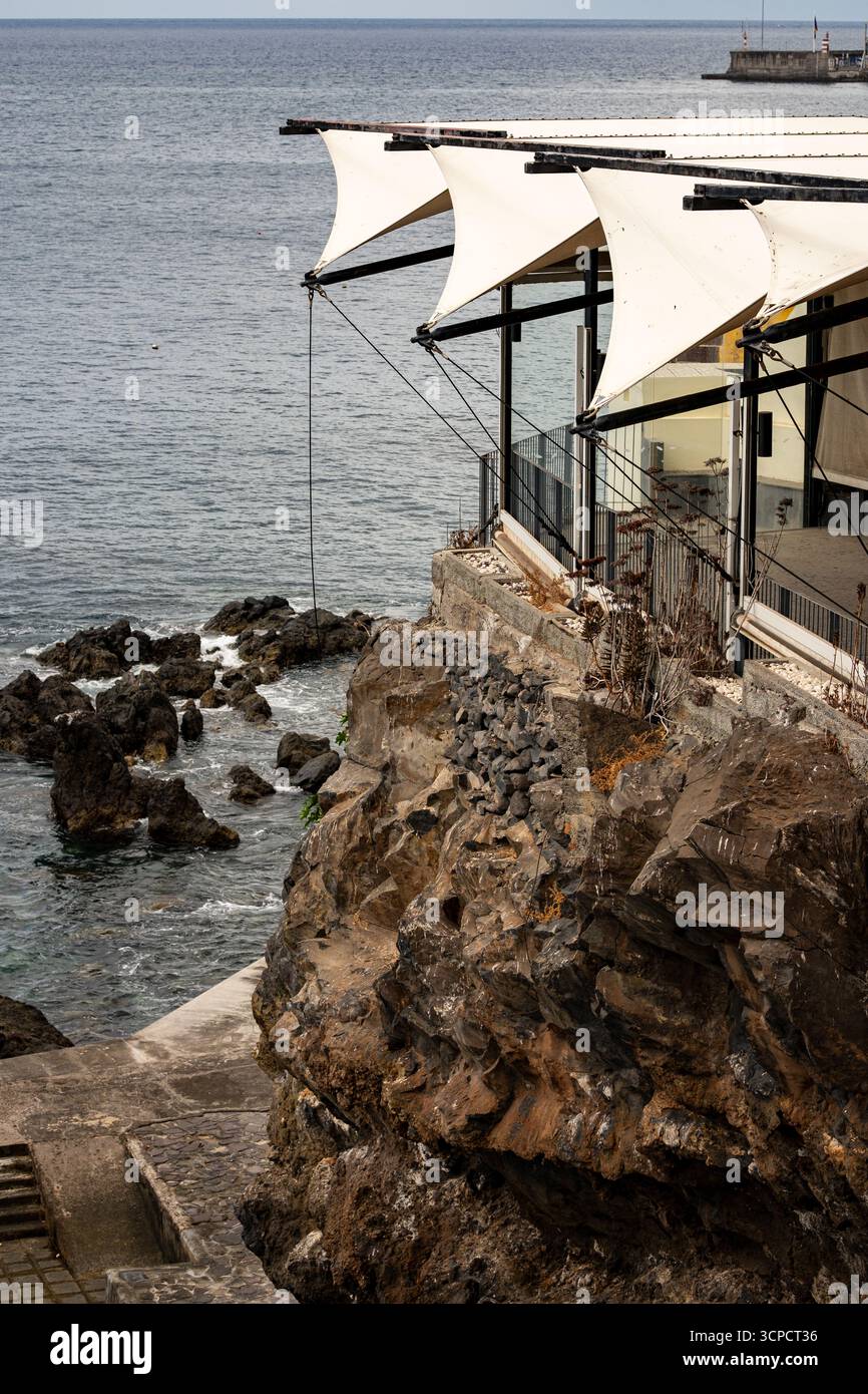 Ein modernes Restaurant oder Café mit markanten weißen segelähnlichen Markisen ist in eine zerklüftete vulkanische Klippe mit Blick auf den Atlantischen Ozean eingebaut. Stockfoto