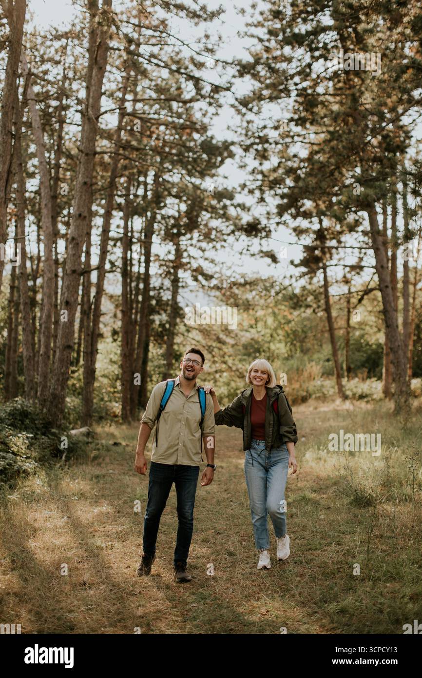 Ein fröhliches Paar schlendert Hand in Hand auf einem sonnendurchfluteten Waldweg, umgeben von hohen Bäumen und üppigem Grün. Sie genießen einen entspannenden Moment im Freien Stockfoto