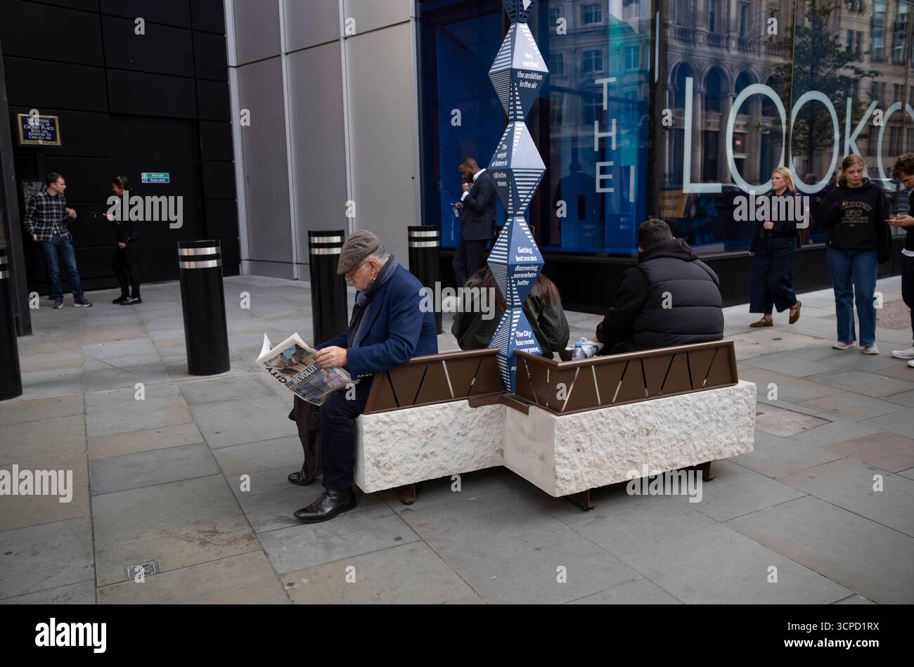 Ein älterer Mann liest eine Zeitung, während er auf einer modernen Bank sitzt, die sich auf dem Bürgersteig am Bishopsgate im Herzen der City of London befindet. Stockfoto