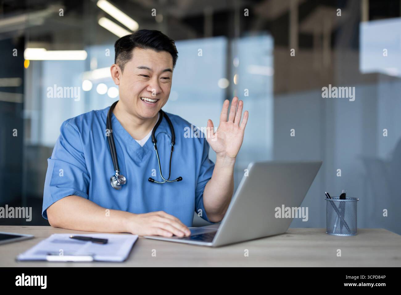 Asiatisch lächelnder junger männlicher Arzt, Student sitzt im Krankenhaus am Schreibtisch in blauer Uniform, grüßt und spricht auf Laptop-Kamera. Stockfoto