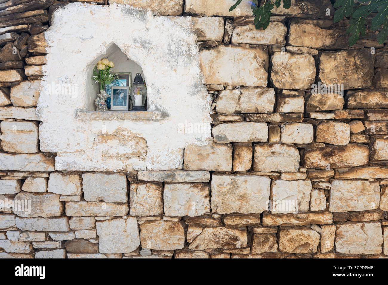Eine geschnitzte Nische in einer verwitterten Steinmauer beherbergt einen kleinen Gedenkschrein mit einem gerahmten Foto, frischen Blumen und einer Kerze, die das Gedenken vermittelt, h Stockfoto