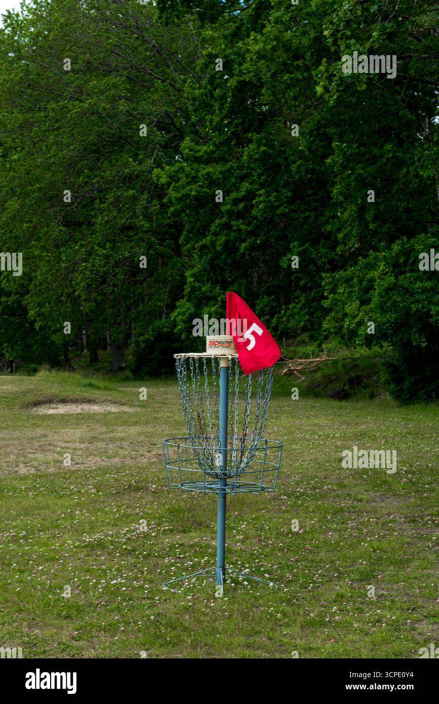 Ein Golfkorb mit Metallscheiben, Ketten und einer roten Flagge, der auf einem grasbewachsenen Feld steht, umgeben von üppig grünen Bäumen in einer Parklandschaft. Stockfoto