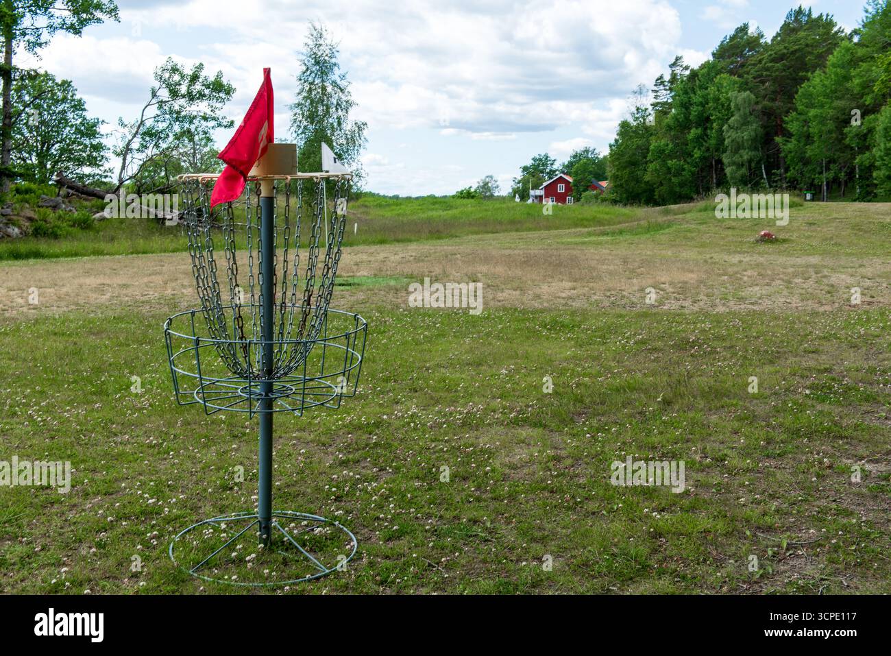 Ein Golfkorb mit Metallscheiben, Ketten und einer roten Flagge, der auf einem grasbewachsenen Feld steht, umgeben von üppig grünen Bäumen in einer Parklandschaft. Stockfoto