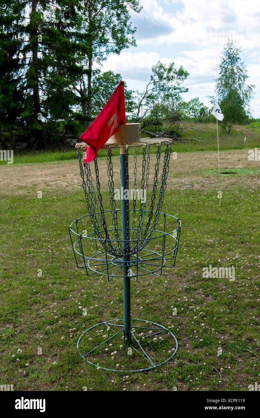 Ein Golfkorb mit Metallscheiben, Ketten und einer roten Flagge, der auf einem grasbewachsenen Feld steht, umgeben von üppig grünen Bäumen in einer Parklandschaft. Stockfoto