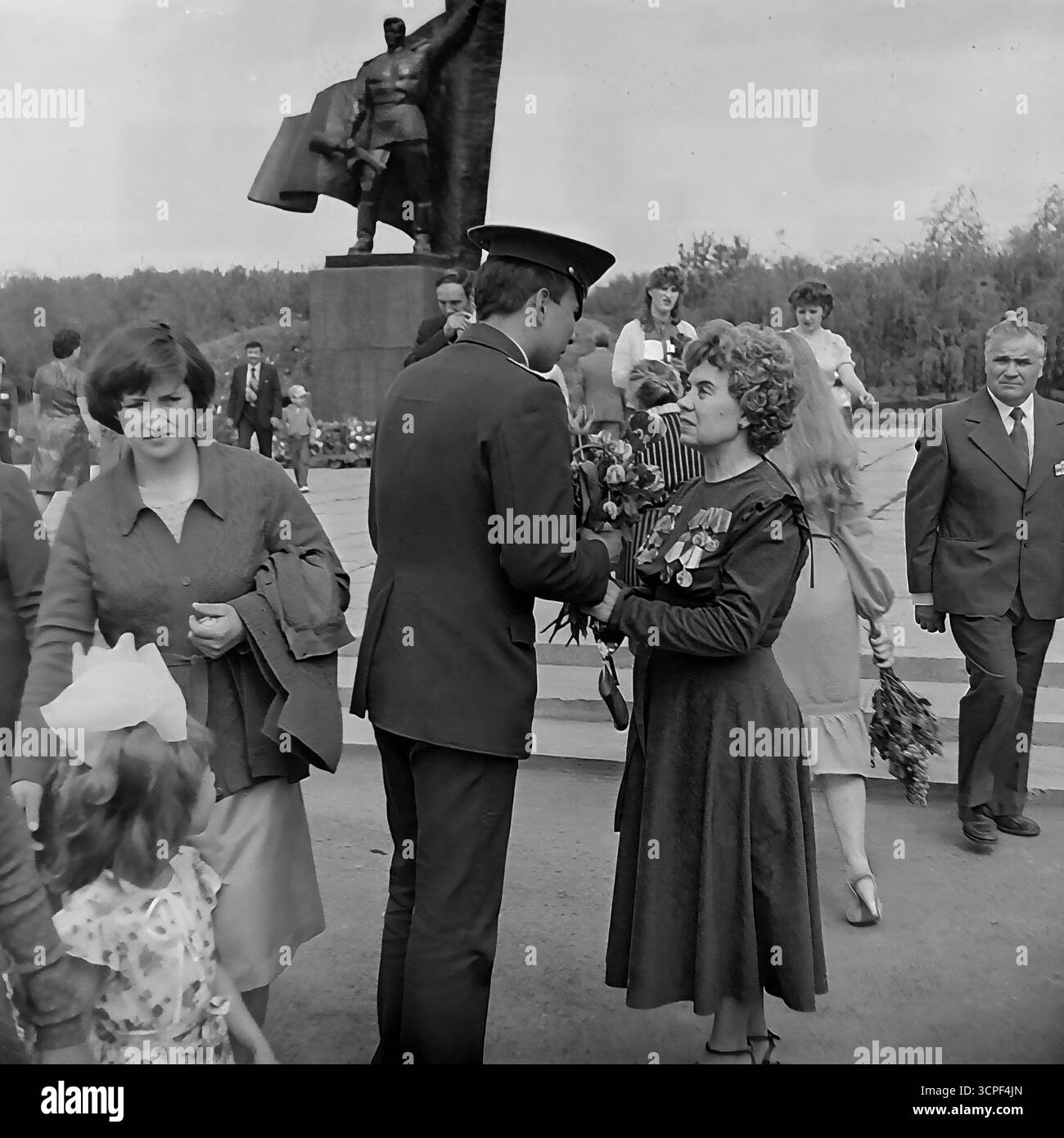 Ein junger sowjetischer Soldat überreicht einer Veteranin aus dem Zweiten Weltkrieg während einer Siegesfeier am 9. Mai in einem Kriegsdenkmal in Slawjansk, der ukrainischen SSR, in den 1970er Jahren Blumen Dieses ergreifende monochrome Bild, das im Lenin-Park (heute Shovkovychnyi-Park) aufgenommen wurde, zeigt ein wichtiges Ritual des Feiertags: Den Akt des Respekts der aktuellen Generation der Armee gegenüber den Veteranen, die im Krieg kämpften. Die Szene symbolisiert die Kontinuität der Generationen, einen Eckpfeiler der sowjetischen patriotischen Ideologie Stockfoto