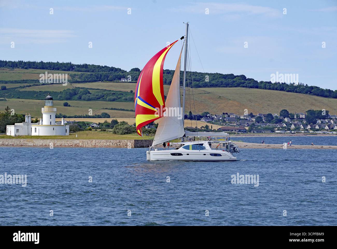 Ein Segelboot mit einem farbigen Segel segelt in der Nähe eines Leuchtturms vor einer grünen Landschaft, Moray Firth, Inverness, Schottland, Großbritannien Stockfoto