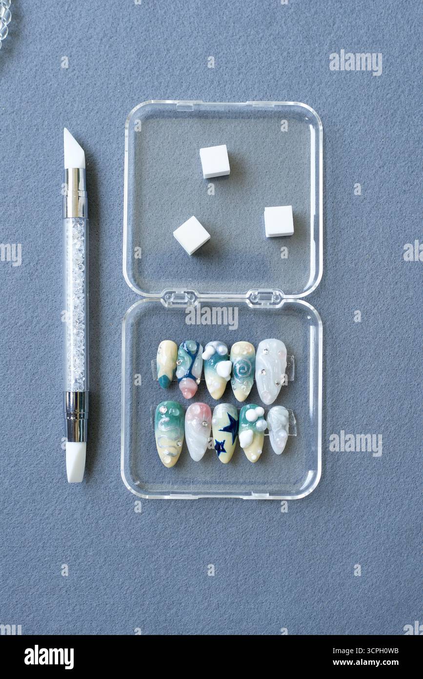 Acrylnagel in zarten Pastelltönen mit Perlen-, Stern- und Wirbel-Verzierungen. Wird in einem durchsichtigen Kasten mit Nagelwerkzeug und Schwamm auf grauem Hintergrund angezeigt Stockfoto