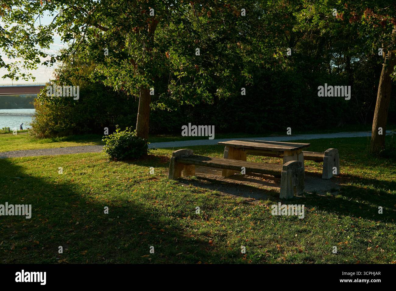 Hölzerner Picknicktisch mit Bänken steht unter warmem Sonnenlicht am Flussufer. Frühherbstszene mit goldenem Gras, ruhigem Wasser und friedlichem natürlichem A Stockfoto