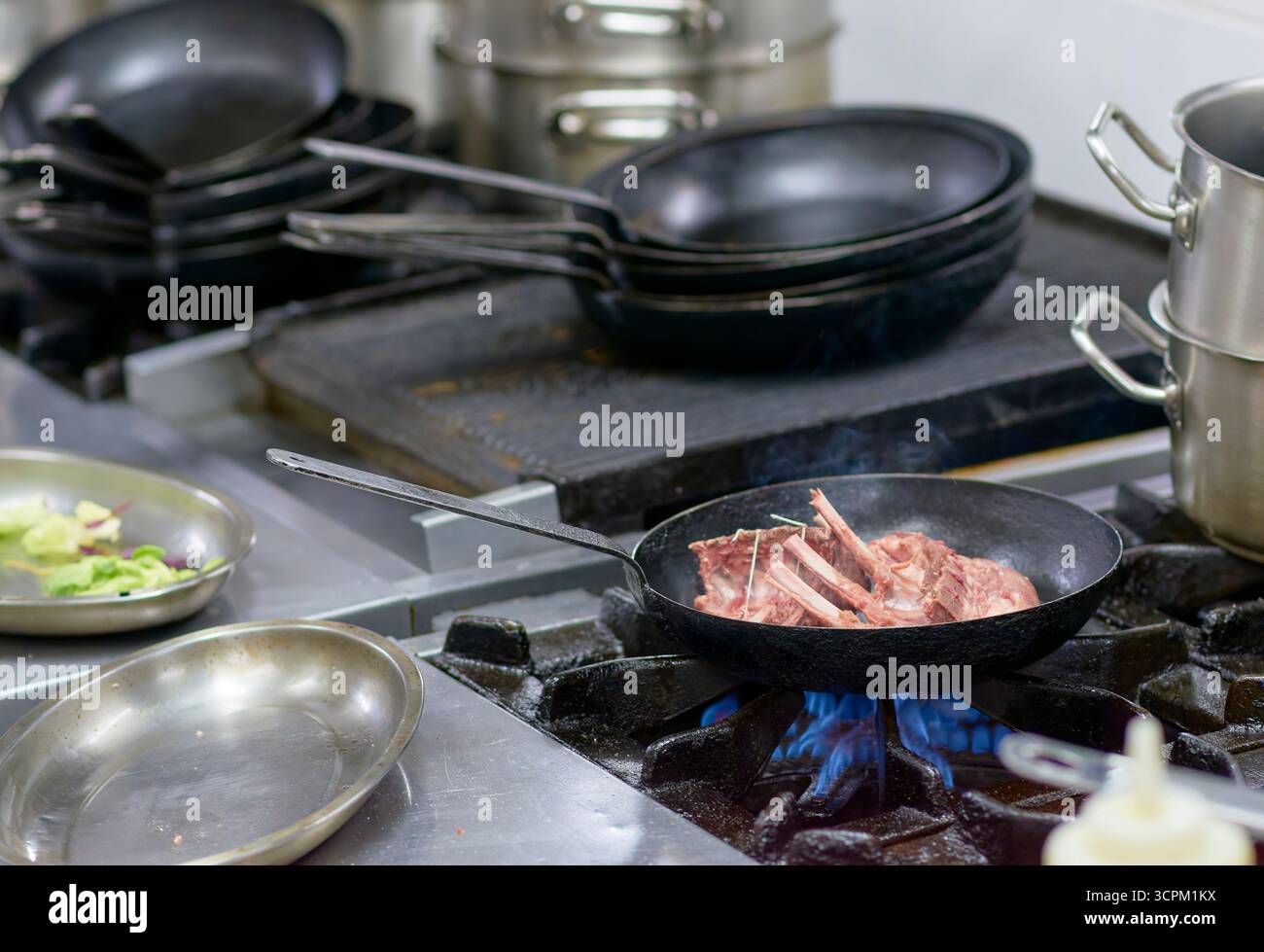 Lammrippen werden in einer gusseisernen Pfanne über blauen Flammen gebraten. Echte Szene in einer professionellen Küche mit Metallutensilien und sichtbarem Dampf. Stockfoto