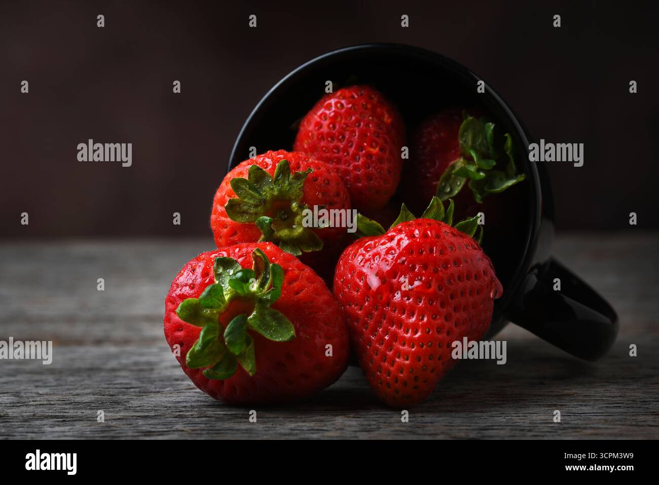 Erdbeerstillleben. Ein schwarzer Becher auf der Seite mit frisch gepflückten Erdbeeren, die auf einem rustikalen Holztisch verschüttet werden. Stockfoto