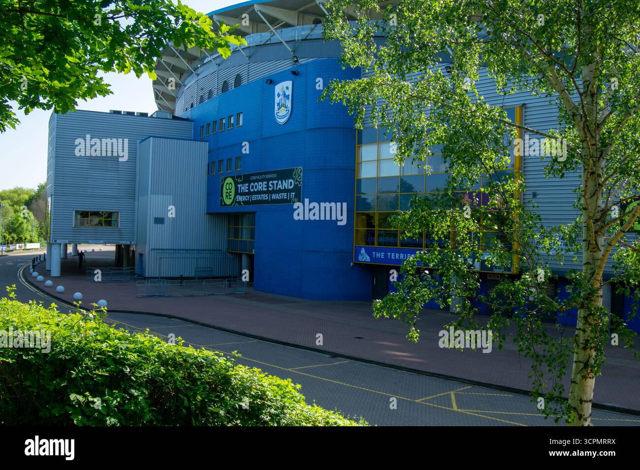 Ein Seitenblick auf das Kirklees-Stadion in Huddersfield mit seiner blau-silbernen Fassade, die an einem sonnigen Tag teilweise von grünen Bäumen eingerahmt ist. Stockfoto