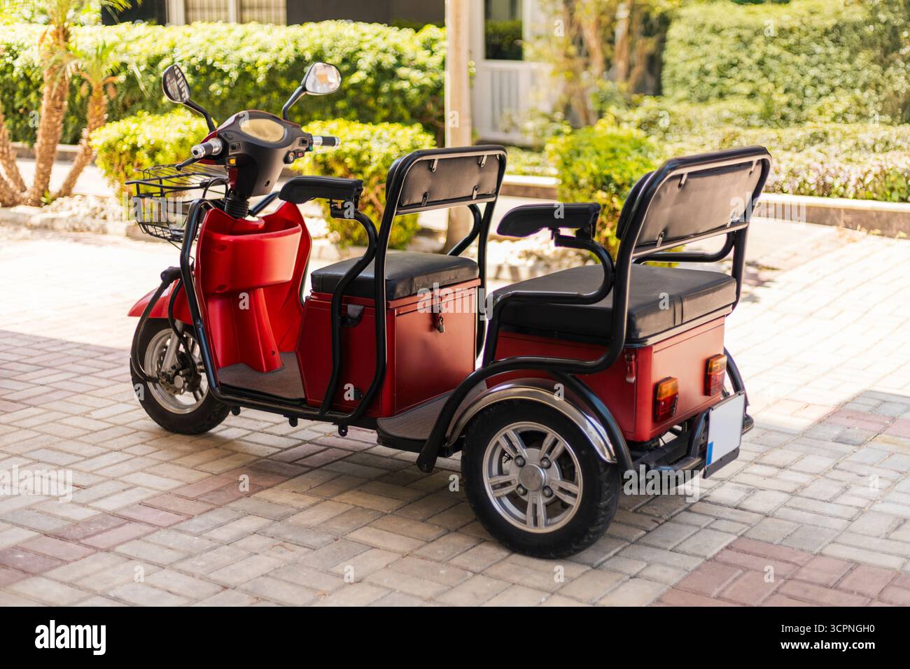 Ein roter Elektroroller mit Beiwagen parkt auf einem Steinweg in einem sonnigen Garten. Der Roller verfügt über zwei Sitze, einen Frachtkasten und üppiges Grün Stockfoto