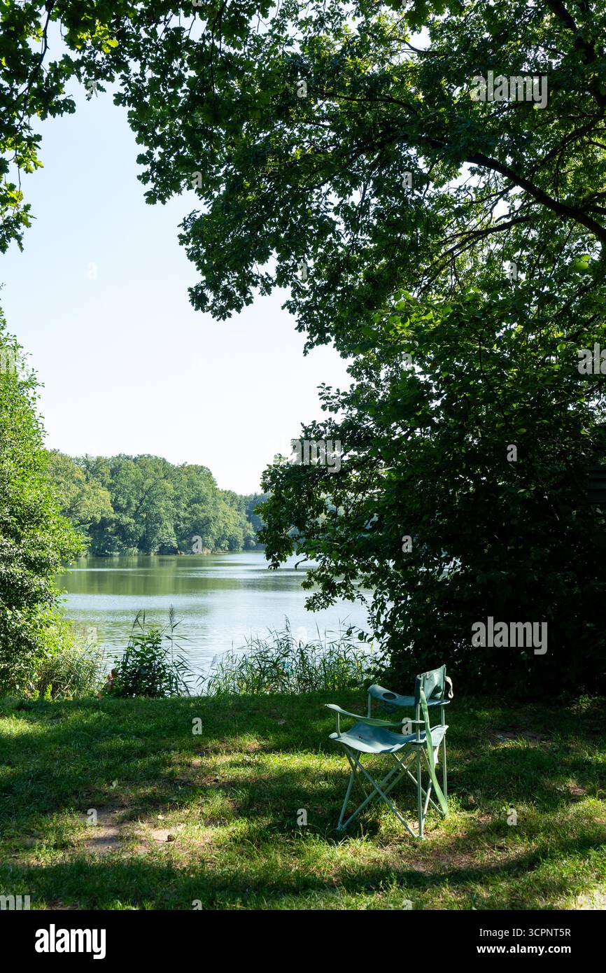 Ruhiger Campingplatz am See mit Klappstuhl unter grünem Baumschatten mit Blick auf das ruhige Wasser und die Waldküste. Konzept der Entspannung im Freien, n Stockfoto