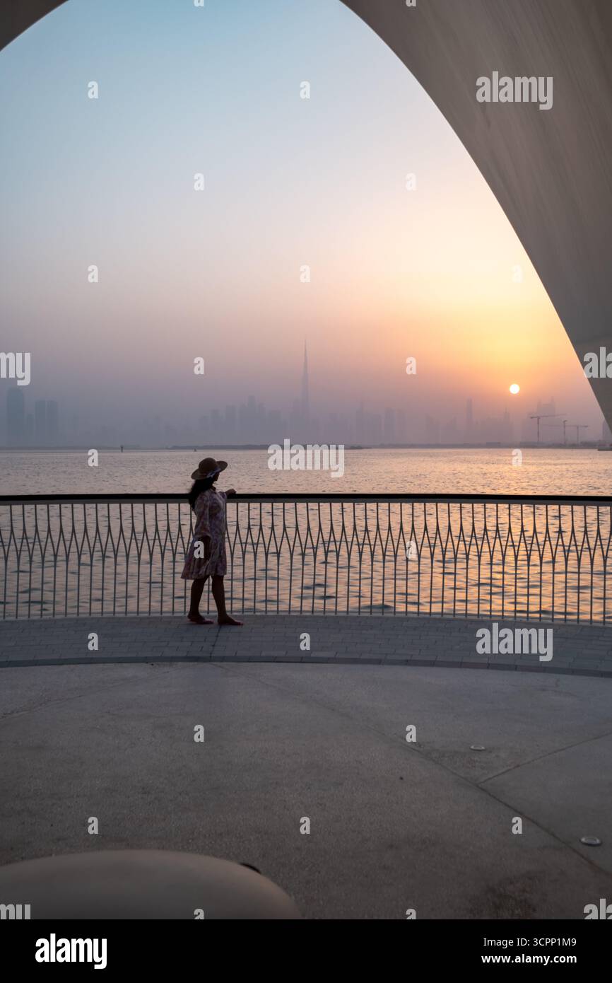Malerischer Sonnenuntergang am Hafen von Dubai Creek hinter der Skyline mit einer jungen Frau, die Touristen spazieren geht, vertikales Stockfoto Stockfoto