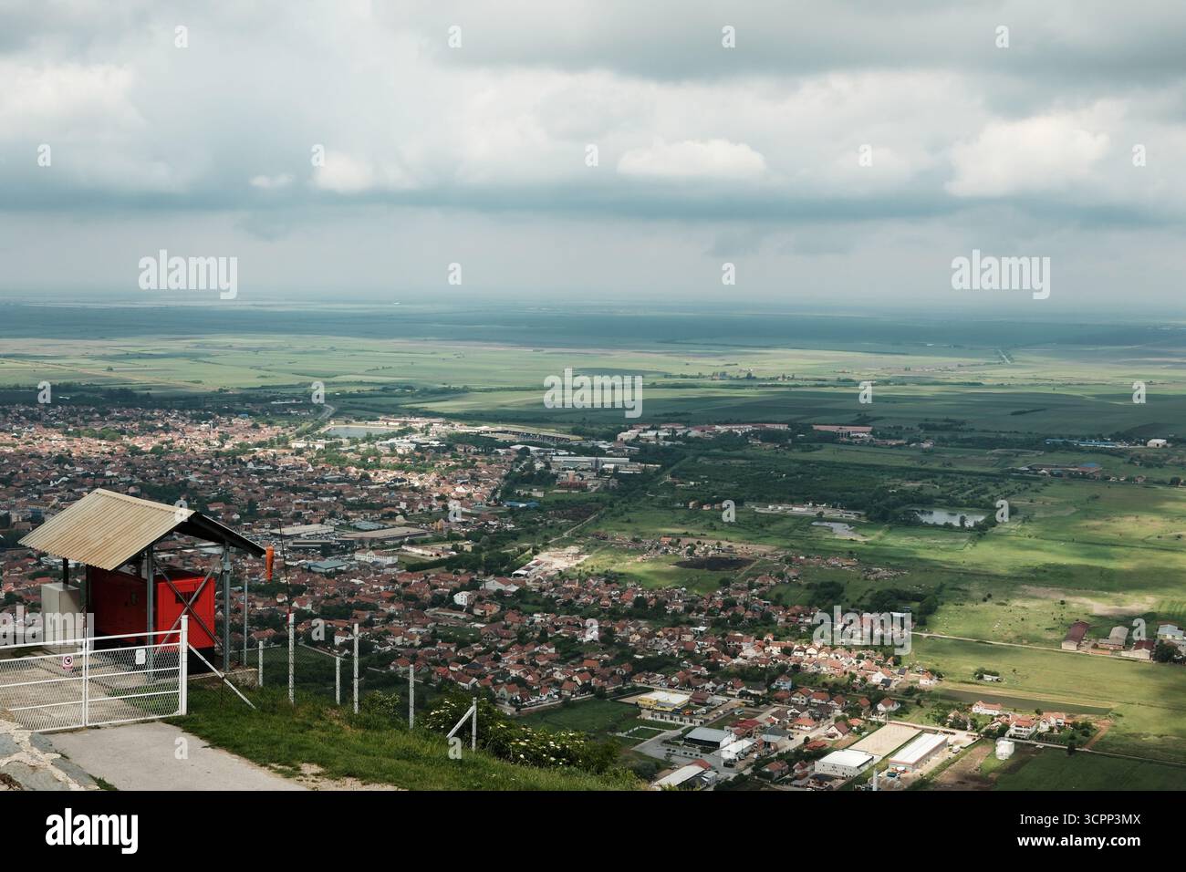 Malerischer Blick auf die Stadt Vrsac mit bewölktem Himmel und umliegendem grünem Ackerland, von einem Hügel aus gesehen. In der Ferne können Sie die rot gekachelten Dächer sehen. Stockfoto