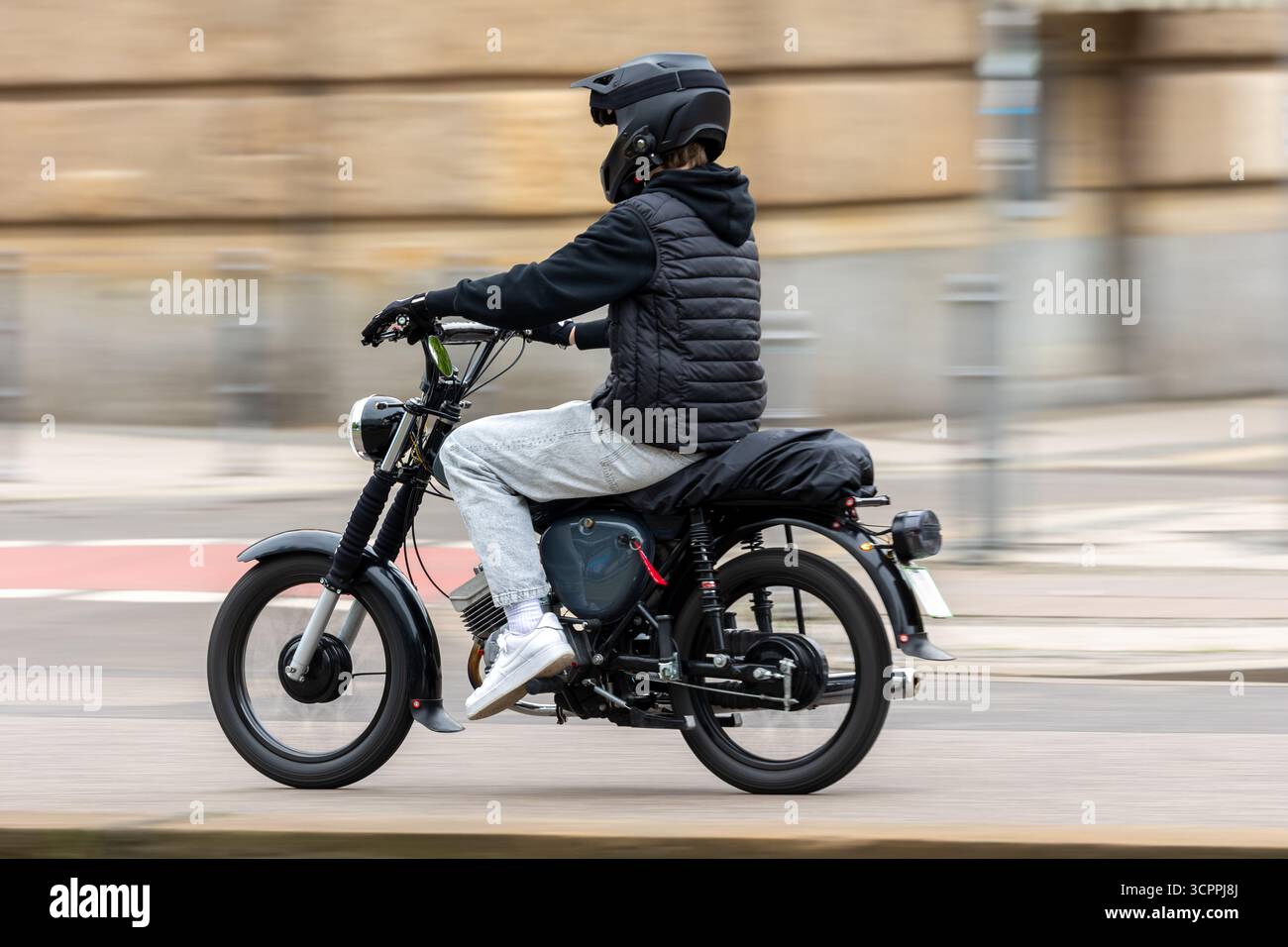 Junge Motorradfahrer in lässiger Kleidung und Helm fahren mit schwarzem Motorrad auf der Stadtstraße. Dynamische Aufnahme mit Bewegungsunschärfe betont Geschwindigkeit, Energie und Stockfoto