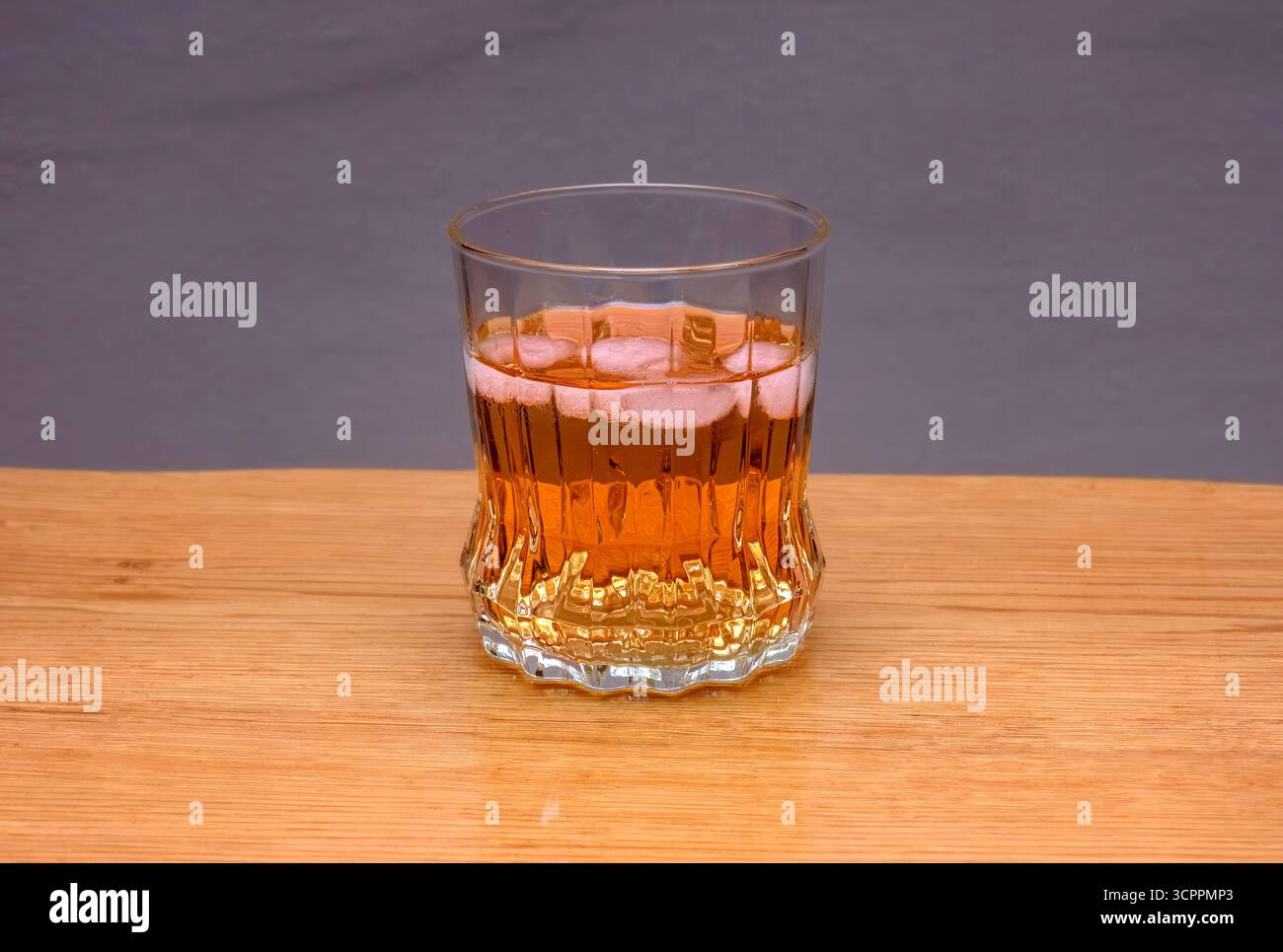 Ein klares Glas, gefüllt mit einer bernsteinfarbenen Flüssigkeit und Eiswürfeln, steht auf einem hellen Holztisch vor einem neutralen grauen Hintergrund. Stockfoto