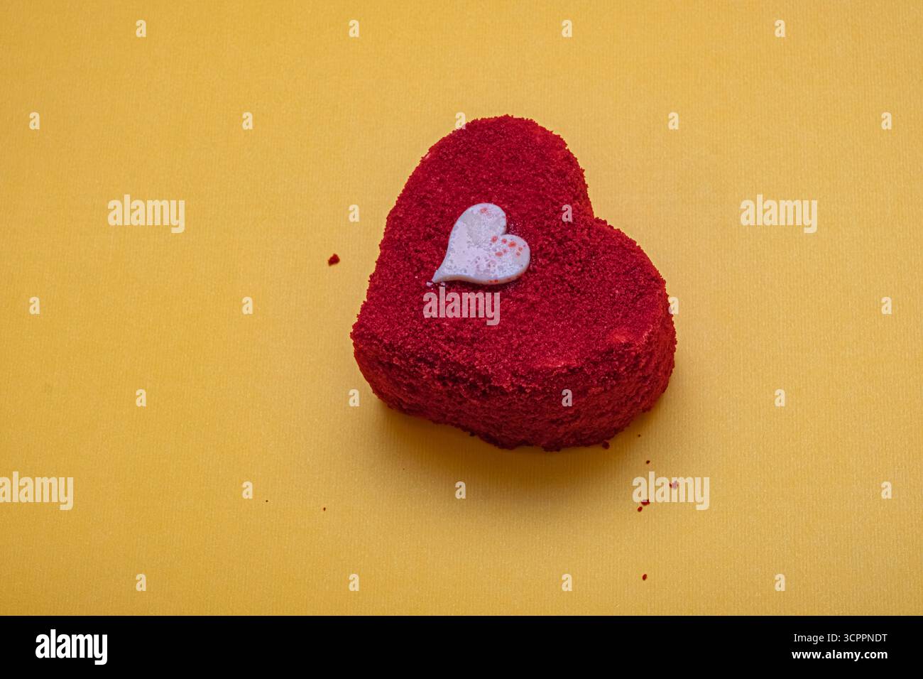 Wunderschön gefertigter roter Samtkuchen in Herzform auf strukturierter Oberfläche. Kleines, dekoratives weißes Herz auf der Oberseite. Valentinstag, Romantik und süßer Genuss. Stockfoto