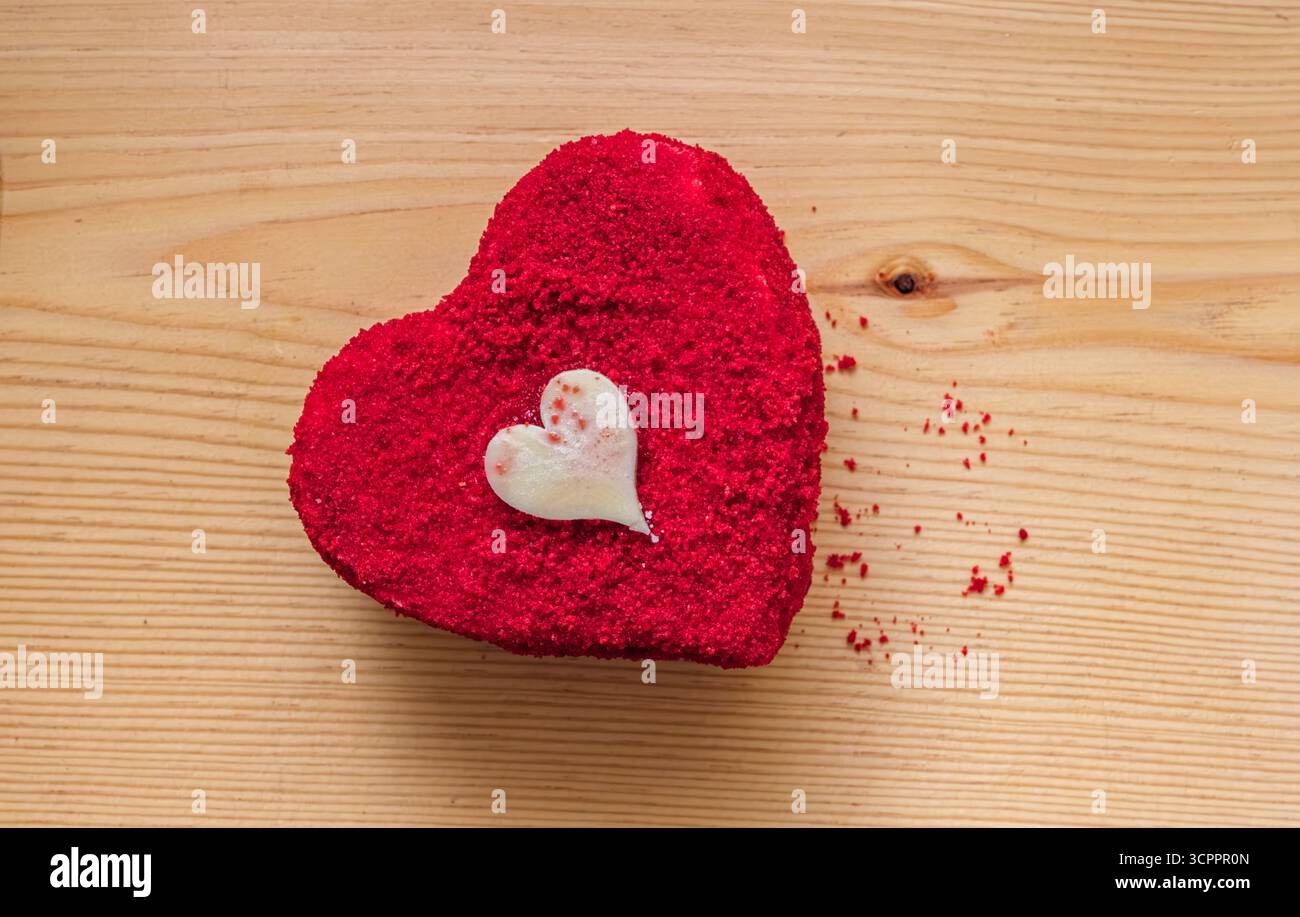 Herzförmiger roter Samtkuchen mit weißem Schokoladenherz auf einer Holzoberfläche. Krümel verstreut für eine rustikale, romantische Note. Stockfoto