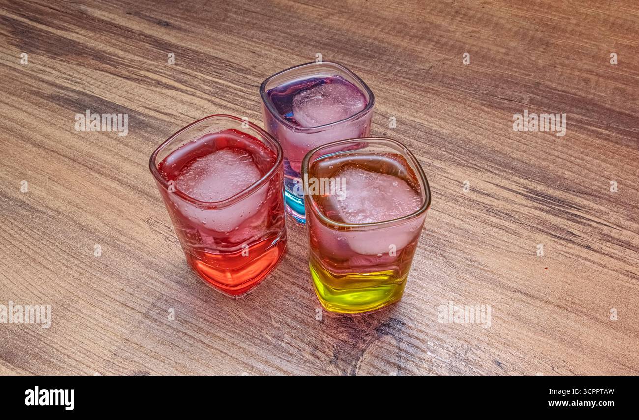 Drei bunte Cocktails. Drei lebendige Cocktails in quadratischen Gläsern mit Eiswürfeln auf Holzoberfläche. Für das Nachtleben und gesellschaftliche Zusammenkünfte. Stockfoto