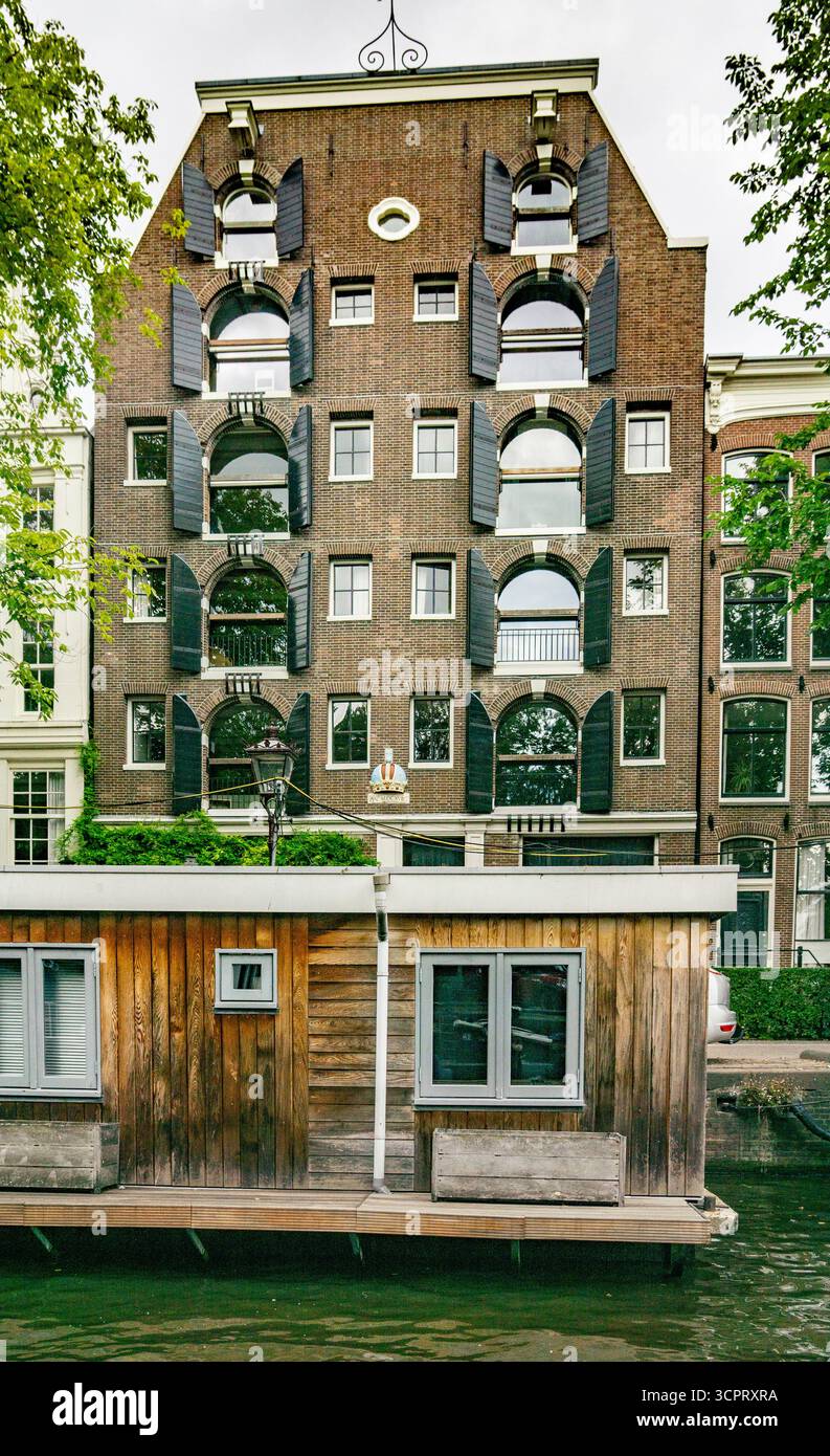 Ikonisches Haus= mit schwarzen Fensterläden auf einem Kanal mit einem Hausboot in Amsterdam, Niederlande Stockfoto